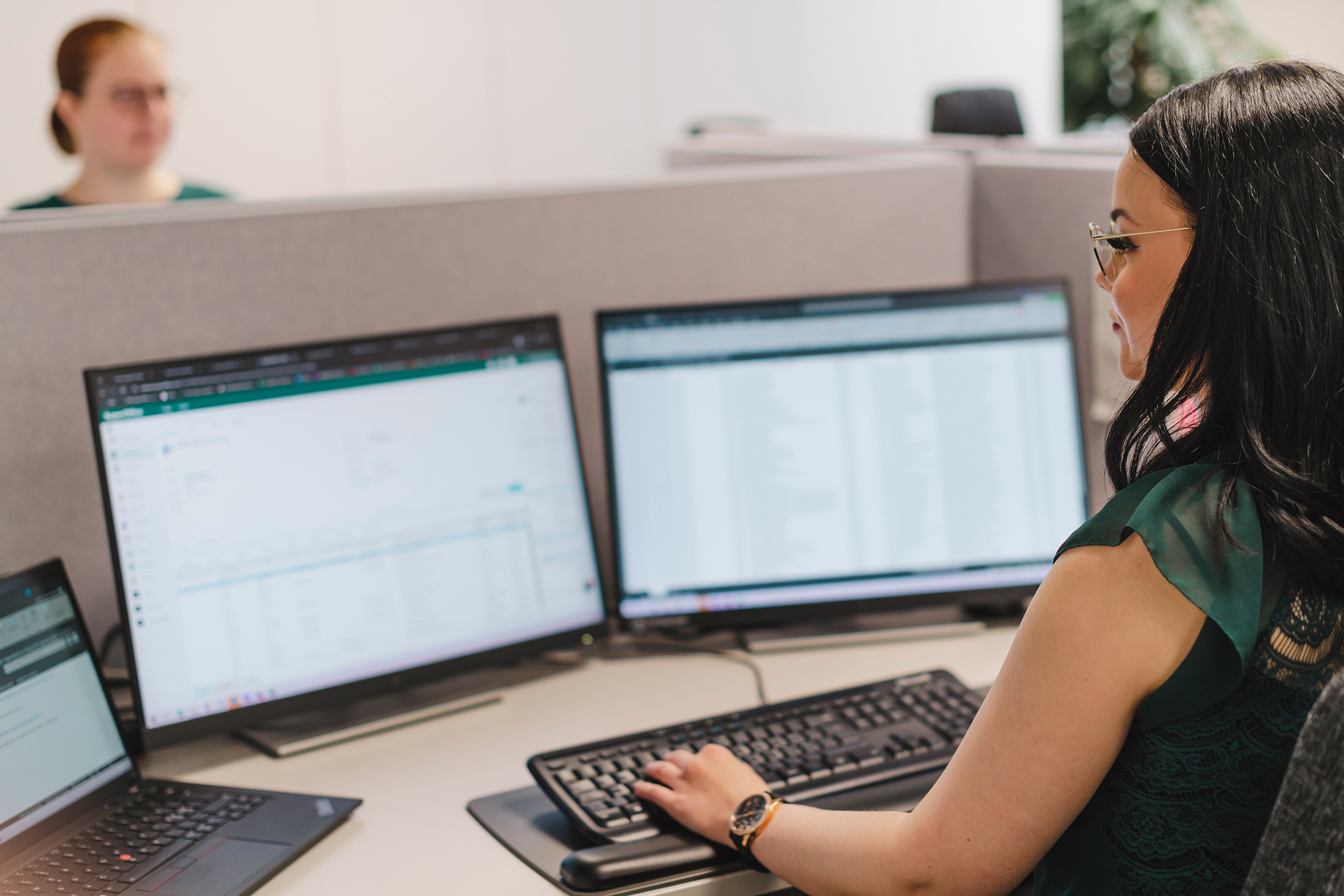 Back/profile view of female employee working, with a glimpse of a female employee across from her behind the screens and a desk divider