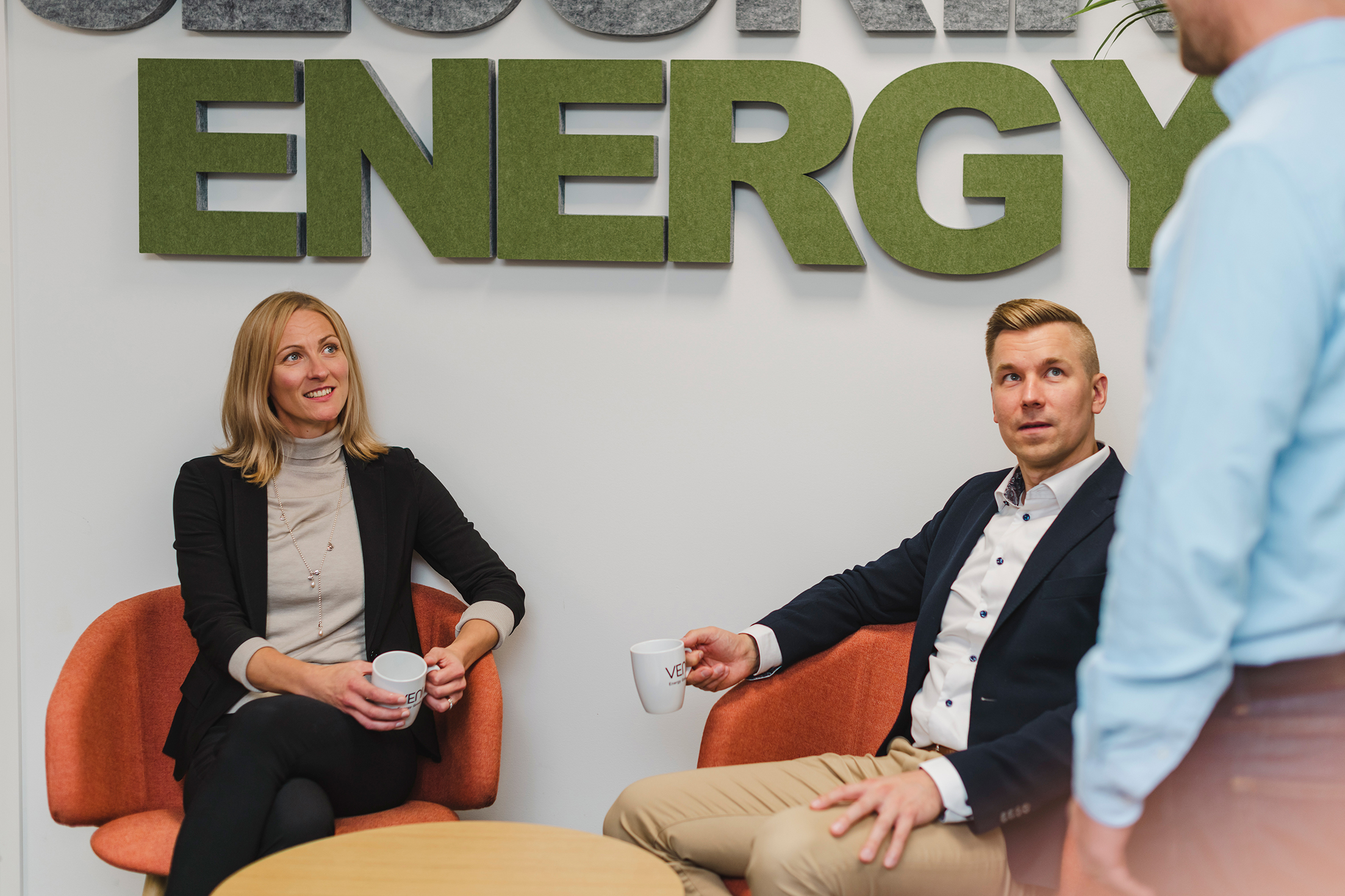 Employees Minna Lindholm and Arto Peltola sitting down in front of sign on the wall reading "energy" and having a chat with an employee entering the frame from the right