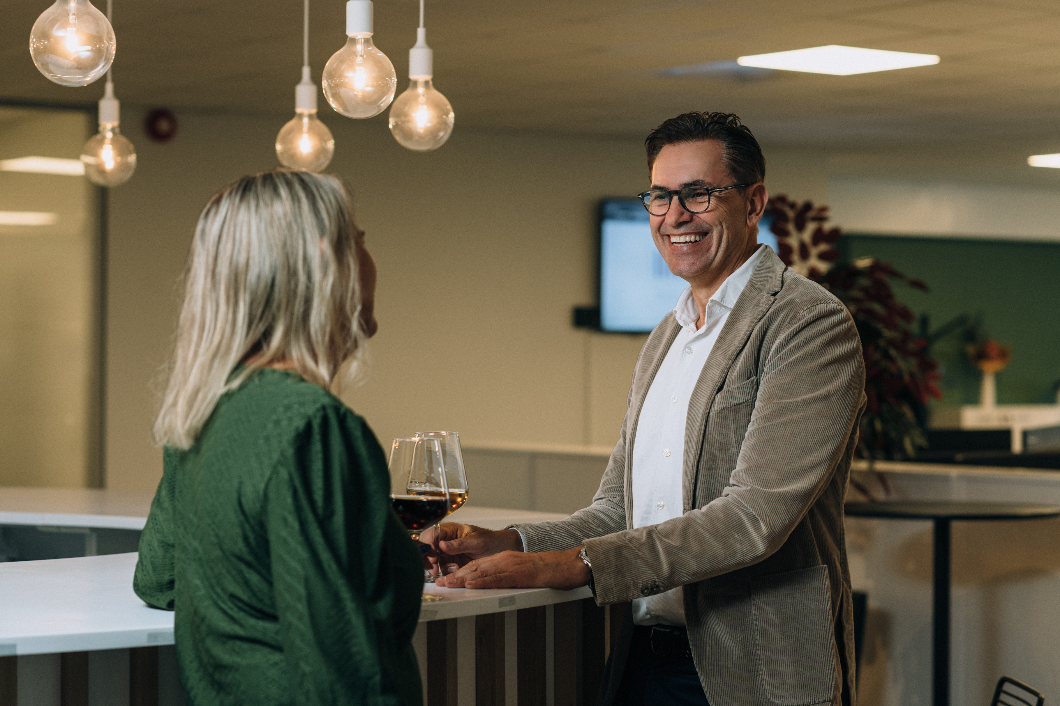 A female employee seen from the back speaking to employee Trond Lutdal in a bar setting with hanging lightbulbs overhead